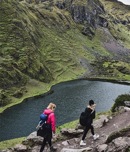 Two hikers with backpacks walk a rocky path beside the dark water of Kinsa Cocha, below steep green slopes