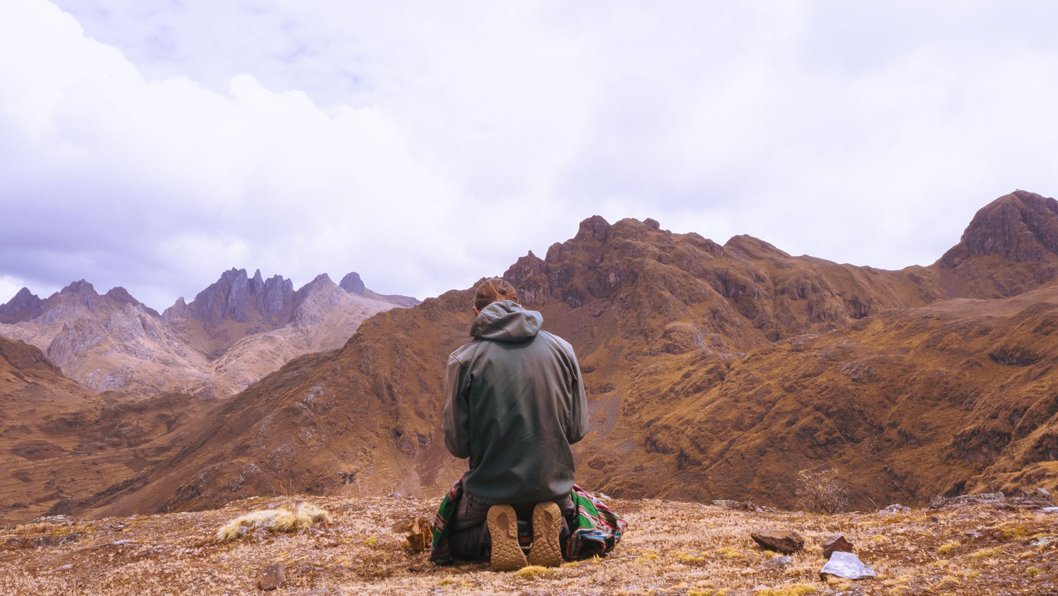 A person in a hooded jacket kneels on a woven cloth, seen from behind, facing rocky brown mountains under cloud