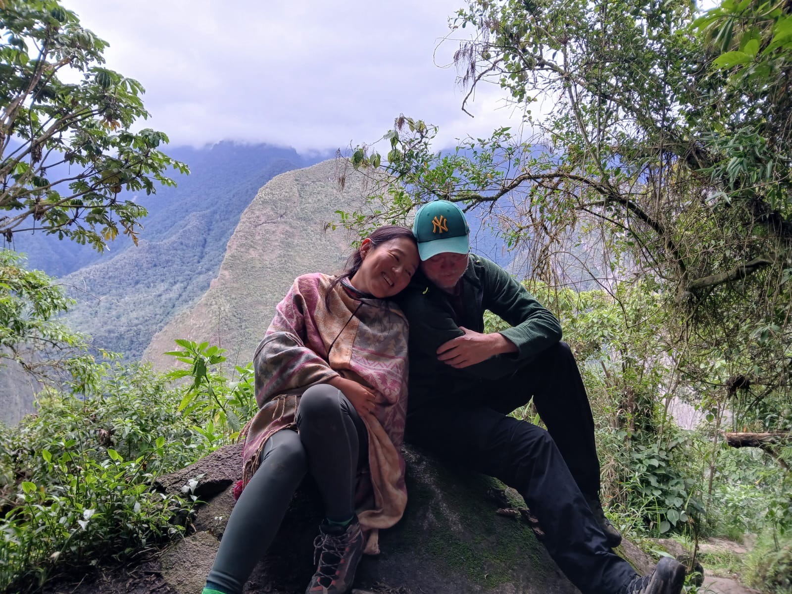 A smiling woman in a patterned shawl leans on a man in a green cap, both seated on a rock above a forested valley