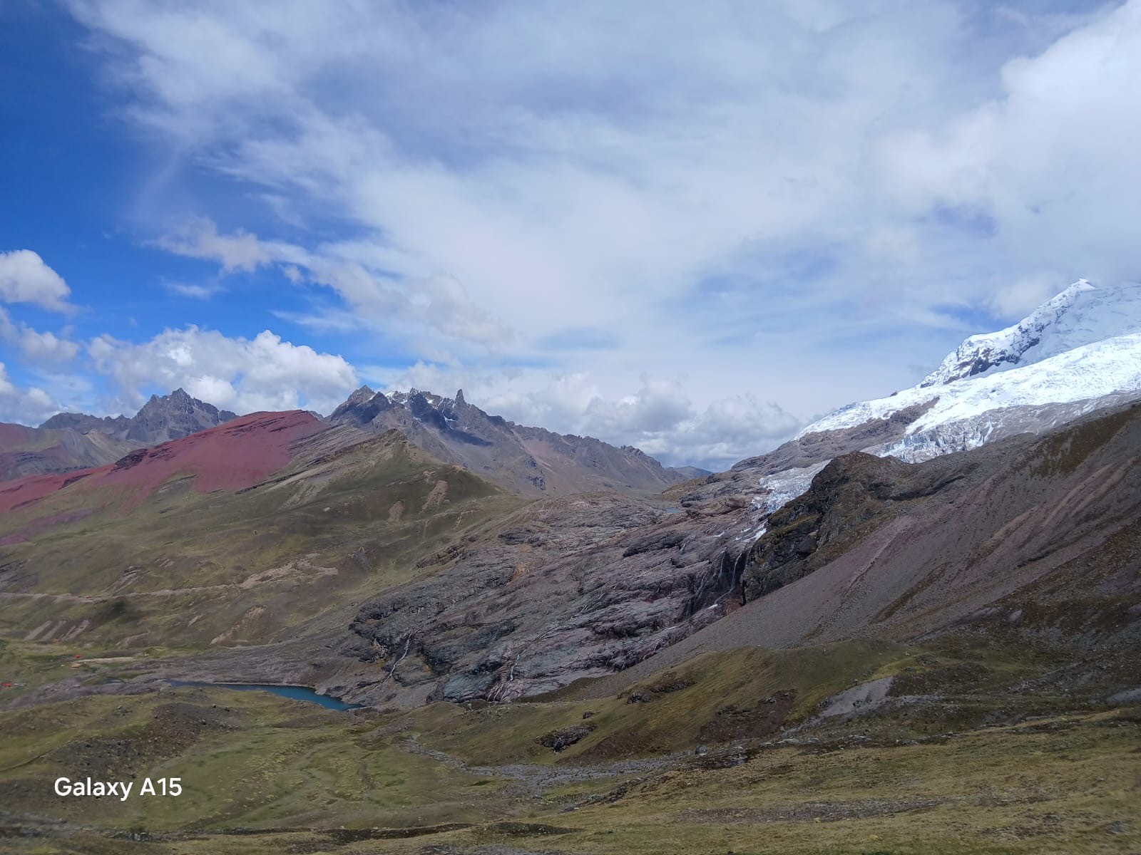 A high mountain valley with a red hillside, a glacier on a snowy peak, waterfalls over dark rock and a small blue lake