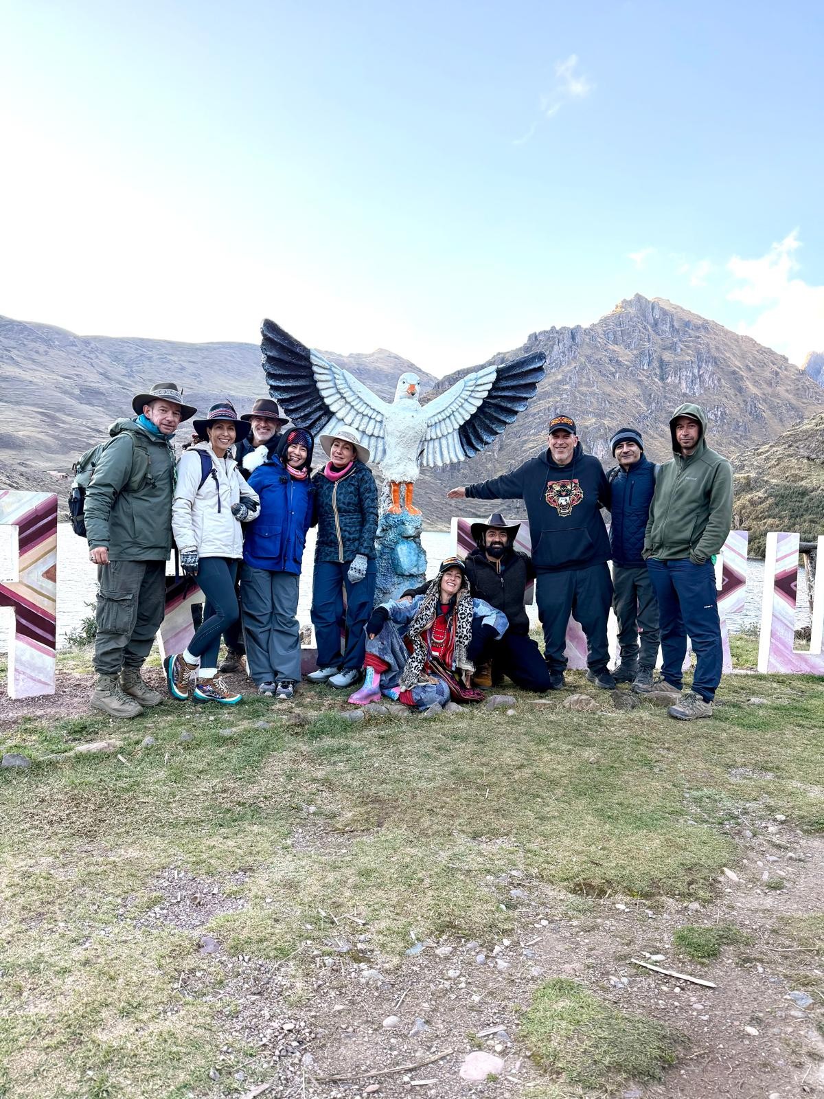 Ten people in hats and warm jackets pose by a statue of a bird with spread wings, a lake and mountains behind them